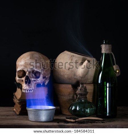 Still life with human skull on old books, smoke on clay pots and blue flame up from metal bowl