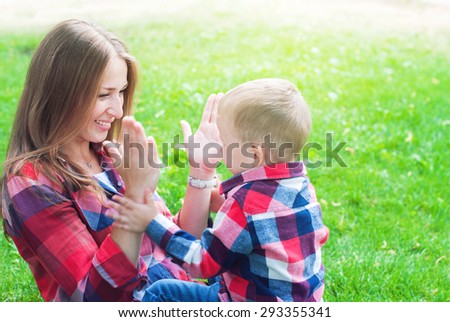 Happy Mother and Her Son in Similar Shirts and Jeans Playing in Hands. They are Sitting on Green Grass in the Summer Park