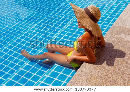 Young woman in a yellow swimsuit and a hat seating down in a swimming pool and having a sun bath on a sunny summer day