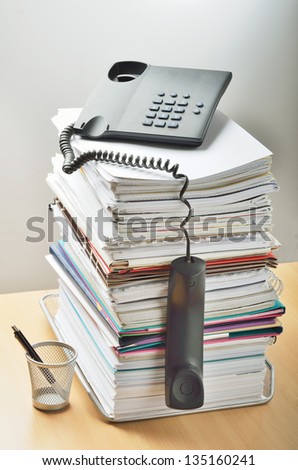 A huge pile of paperwork on a desk and a phone above it