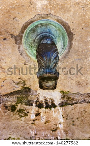 Antique marble water spout, colored bronze and green, on neutral colored, textured marble wall with water droplets flowing from spout.