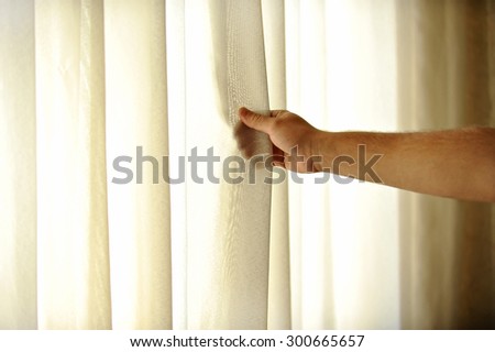 A man\'s hand pulling a window curtain for warm daylight to enter the room