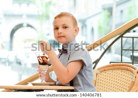 A boy in a street cafe eating ice cream. A handsome boy is sitting in a chair on a city street cafe eating ice cream of a large vase. Eating ice cream with a spoon. The boy in the shirt.