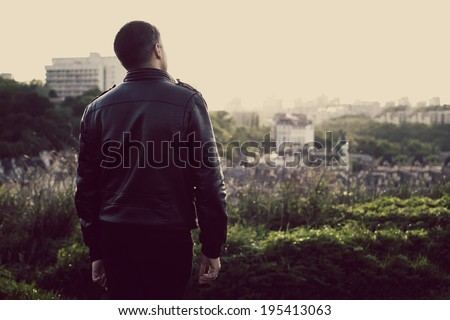 Modern adult sad man turned back. Man on the background of the city and nature at sunset. A man in a black jacket s skin looks and thinks about something.