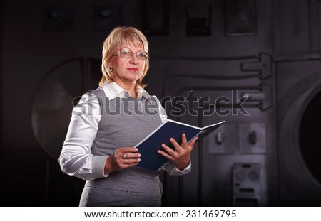 Adult business woman. Woman engineer by profession designer. She is a scientist or researcher. Woman with a book for records worth on the dark background wall of an industrial building.