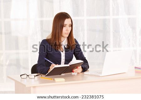 Woman student or beginner office staff at work in the office. She works at his laptop beech and makes notes in a notebook. The girl secretary or office manager sitting at the table on time of the day.