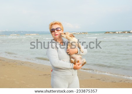Adult woman is holding her dog. Retired woman standing on the shore of the sea or ocean. Old woman spends time with his dog on the background of the sea. Walking the dog on the beach. Tourism. Dog.
