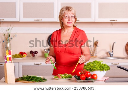 Smiling old housewife in modern kitchen. Adult woman preparing a salad in the kitchen interior. Woman making healthy food standing happy smiling. She in kitchen preparing vegetable salad at home.