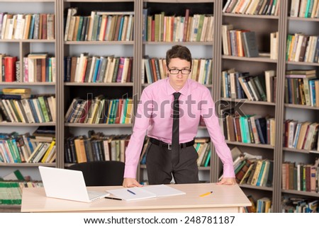 Office staff and student intern working at the table on the background of shelves with books. Work in the library or in the archive. Young man near the desk with a laptop and a notebook to take notes