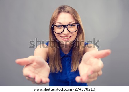 Girl in glasses. Hands in front. Young beautiful modern woman. Various women\'s emotions. Portrait of attractive cheerful young woman in glasses pointing at space over gray background, toned.