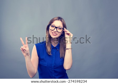 Girl in glasses show victory sign. Young beautiful modern woman. Various women\'s emotions. Portrait of attractive cheerful young woman in glasses pointing at space over gray background, toned.