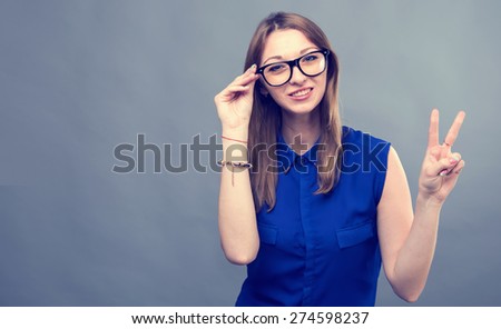 Girl in glasses show victory sign. Young beautiful modern woman. Various women\'s emotions. Portrait of attractive cheerful young woman in glasses pointing at space over gray background, toned.