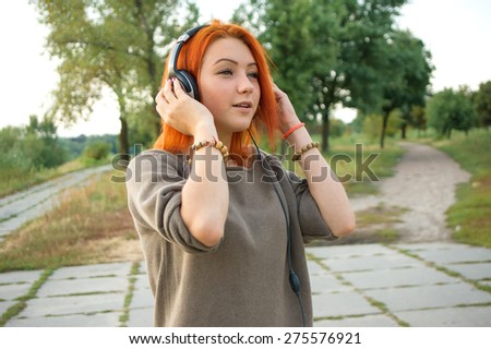 Young and beautiful woman wearing headphones listening to music and singing on the background of nature city park. Woman with headphones listening music. Music teenager girl dancing against.
