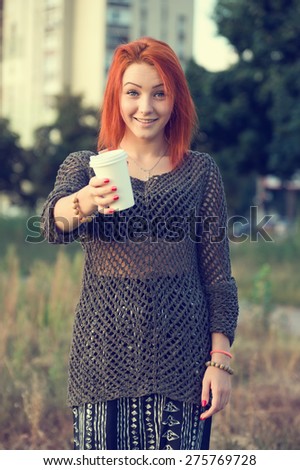 Red hair woman drinking tea or coffee from a white paper disposable cup. Concept - tea or coffee cup. Stretching forward with coffee cup. Young beautiful girl on the background of nature city park.
