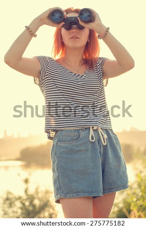 Girl with binoculars at the beach or river in the rays of the evening sunset. Young beautiful woman on nature background. She is resting in the open air. Keep holding binoculars. Look ahead.