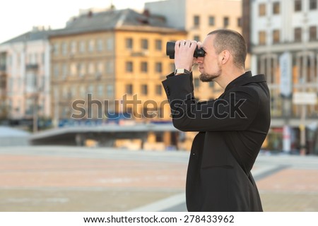 Man and binoculars. Business man with binoculars in his hand against the background of the streets downtown. Security man holding binoculars. Looking for a job. Man with spying. Service monitoring.