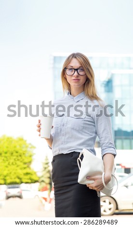 Modern woman office worker wearing glasses. She went out for lunch on the street. Business woman holding a cup of coffee or tea. She is against the background of business office buildings and parking.
