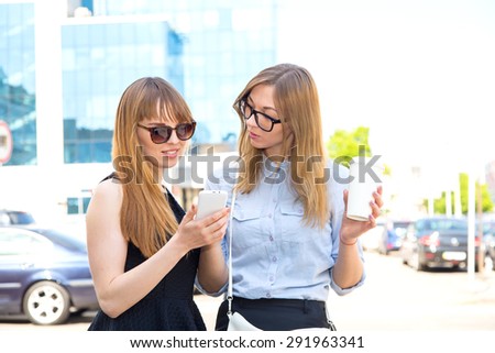 Two happy women friends sharing social media in a smart phone outdoors in a city. Two young women looking at mobile phone together while standing outdoors city. Drink coffee and discuss news. Meeting.