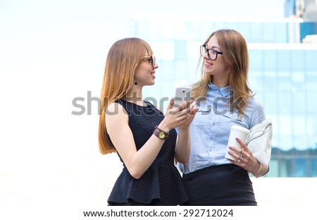 The two business women met while working break. In the first woman in the hands of the mobile phone, from the second girl with a paper cup of coffee. Two young women discuss the news and shopping.