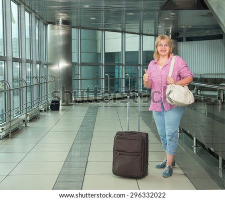 Adult woman is traveling. Woman with suitcase standing in the building bus station or airport. Holding her passport and ticket. Retired woman embarks on a journey with his travel luggage. Thumbs up.