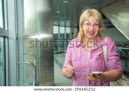 Beautiful adult woman, smart phone. Woman using app trip on a mobile phone. See maps application in the mobile phone. Woman is traveling. Bus station or airport. Thumb up concept. Happy adult woman.