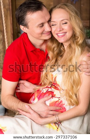 Close up pregnant woman in bright color dress and man sitting on a swing against the backdrop of a country house. Woman and man happiness. Man and beautiful woman is expecting a baby, she is pregnant.