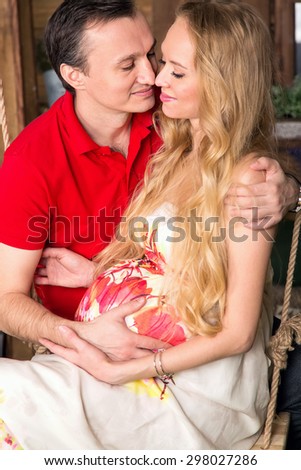 Close up pregnant woman in bright color dress and man sitting on a swing against the backdrop of a country house. Woman and man happiness. Man and beautiful woman is expecting a baby, she is pregnant.