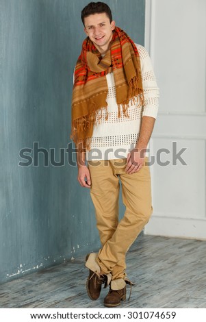 Portrait of an attractive young man - smiling. Young man in white sweater and scarf laughing happily on white blue wall background. Autumn clothes. man in a warm scarf leaning on the wall of the room.