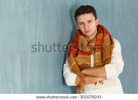 Portrait of a smart serious young man standing against blue background. Young serious man in a white blouse and scarf. Autumn style of dress. A man in a warm scarf leaning on the wall of the room.