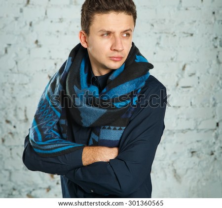 Portrait of a smart serious young man standing against blue background. Young serious man in a blue blouse and scarf. Autumn style of dress. A man in a warm scarf leaning on the wall of the room.
