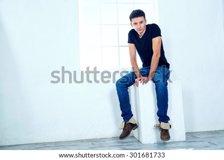 The man next to the window. One man sits with his back to the window. Fashionable male model. The empty space of the room around a man. Against the background of windows and light blue walls.