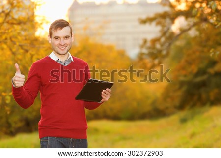 Man with the electronic tablet raised thumbs up. Young man using tablet computer on autumn street and city park. Student using tablet computer. Happy man thumbs up.