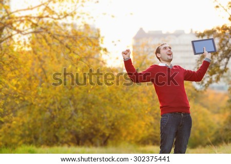 Very cheerful man celebrating victory. Young man using tablet computer on autumn street and city park.  Student using tablet computer. Man winner. Man celebrating, arms raised, expressing positivity.