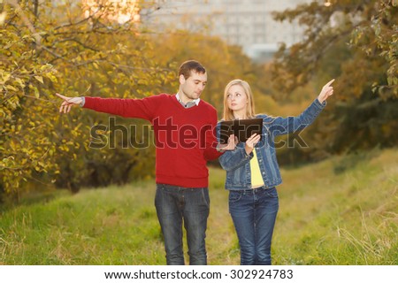Man and woman holding electronic tablet in hands, argue where to go. Point the finger in different directions. Young students. September, October, golden autumn. Traveler, tourist, navigator, lose.