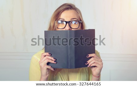 Woman hiding behind the grey book. Woman covering her face. Shy, secret. Woman with glasses hiding face behind book looking at camera surprise. Education concept. Face expression. Girl holding book.