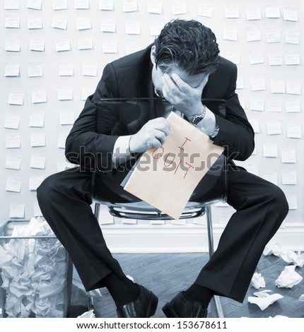Failed stressed businessman - executive concept in blue tone with hands to head facing wall of sticky notes and trash can overflowing with paper holding I Quit sign