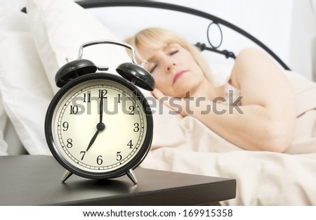 A middle age woman sleeps while alarm clock in foreground reads seven o\'clock.