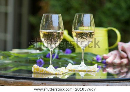 Glass of white wine set outdoors on glass table with flowers and other greenery, as well as a watering can and gardening gloves for a break from gardening