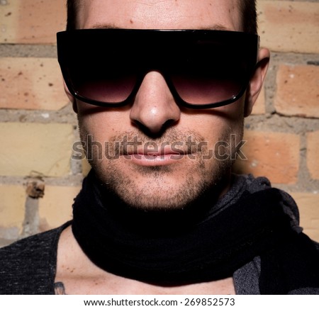 Very tightly cropped portrait of a man with a stubble beard, scarf and sunglasses against brick wall