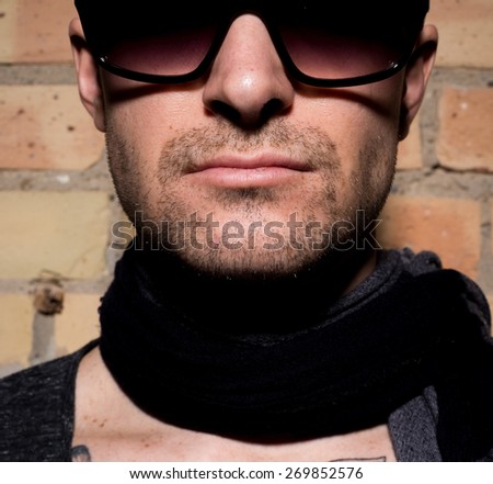 Very tightly cropped portrait of a man with a stubble beard, scarf and sunglasses against brick wall