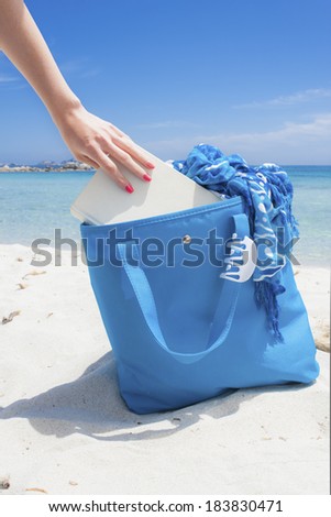 Beach bag on the sand, with book and foulard coming out from it and hair claw hanged on its handle, over girl legs and amazing clear water seascape