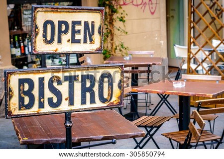 Open bistro sign at the empty cafe terrace, with empty wooden chairs and tables at the outdoor open terrace outside a restaurant or a bar, with some liquor bottles in the window out of the focus