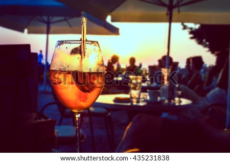 A glass of cold orange cocktail at the sunset on the table of a beach bar at the sunset, with blurry people in the background on a summer evening, with copy space for text. Retro artistic edit.