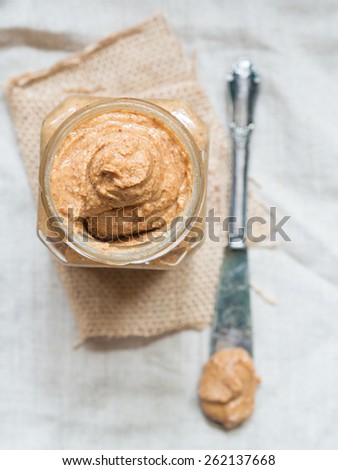 Vertical photo of homemade natural almond butter in a glass jar placed on rustic beige table cloth and with a butter knife on the side. Pastel colors. View from above.