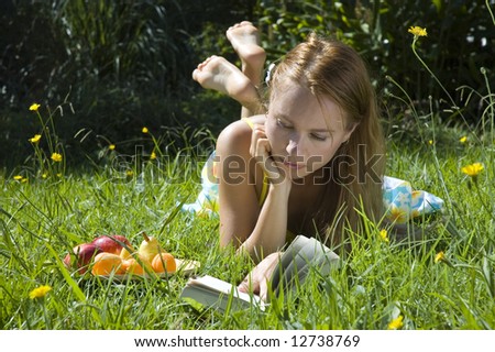 Young blonde woman reading a book on the grass with plate of fruits on summer holidays