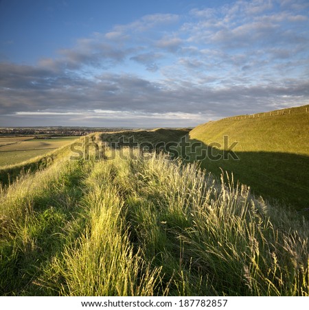 Late evening sunshine rakes across the Iron Age hill fort of Maiden Castle in Dorset, England. Dorchester, the County Town of Dorset, is visible on the horizon.