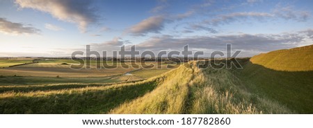 The late evening sunshine rakes across the Iron Age hill fort of Maiden Castle in Dorset, England. Dorchester, the County Town of Dorset, is visible on the horizon.