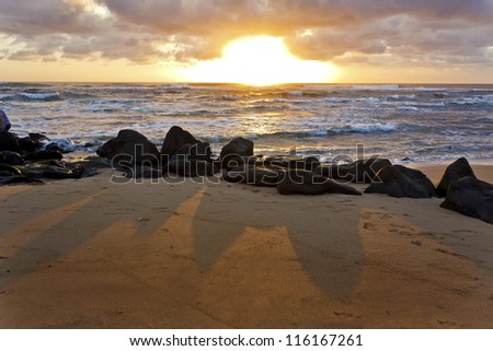 Rocks cast long shadows on the sand as the Sun rises over the Pacific Ocean in Kauai, Hawaii