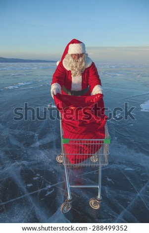 Santa Claus carries a shopping cart with gifts in a sack on a winter lake ice