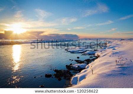 Winter sunset in Iceland. The sun sets into the clouds beyond a river, surrounded by snow covered banks.